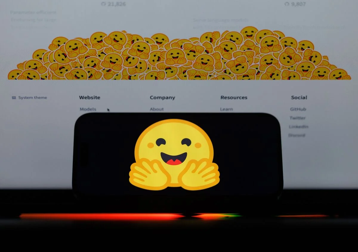 A smartphone showing the Hugging Face emoji logo against a background of the platform website.